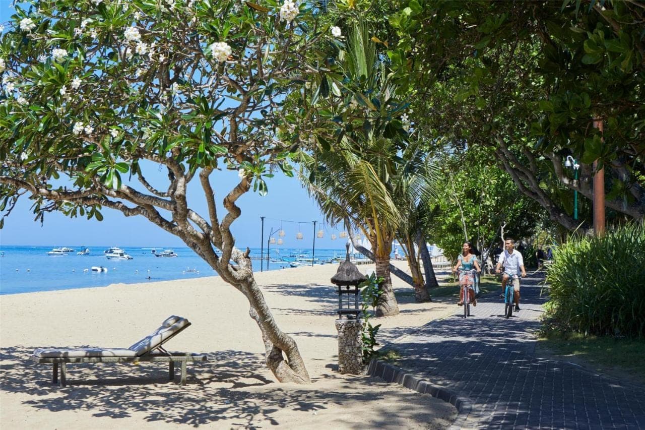 two people bicycling on a beach with a bench and a tree at Hyatt Regency Bali in Sanur
