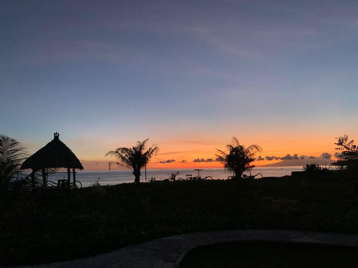 a sunset over the ocean with a gazebo and palm trees at Yogi Beach Cabin in Amed