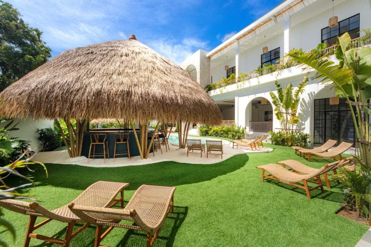 a patio with chairs and a table and a straw umbrella at The Lazy Lama Hostel Uluwatu in Uluwatu