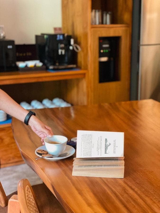 a person reaching for a cup of coffee on a table with a book at Leafy Home Canggu - 5 Mins Walk to Echo Beach in Canggu