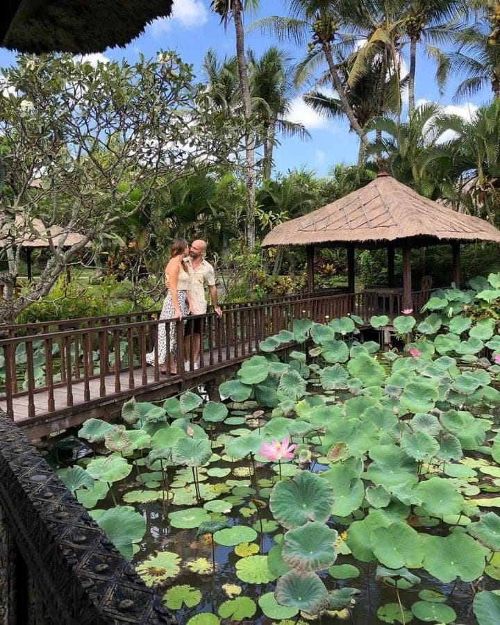 two people standing on a bridge over a pond with lilies at Hotel Tugu Bali, Canggu in Canggu